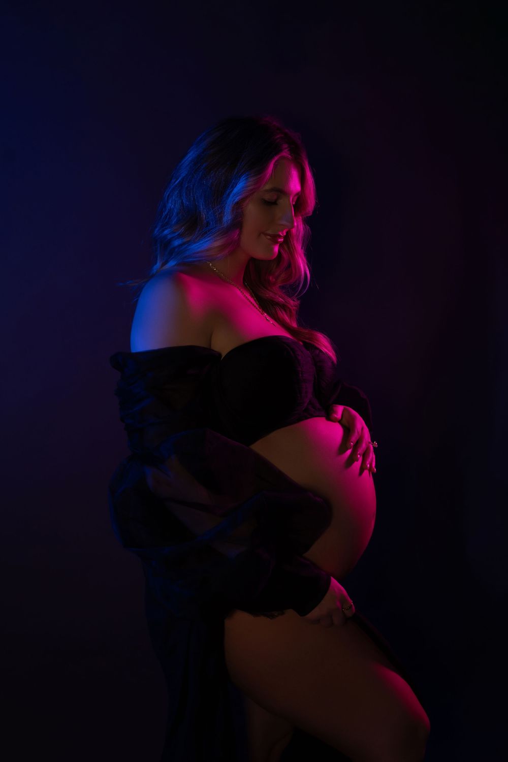 Pregnant woman in black lingerie posing in dramatic pink and blue neon studio lighting, cradling her baby bump.