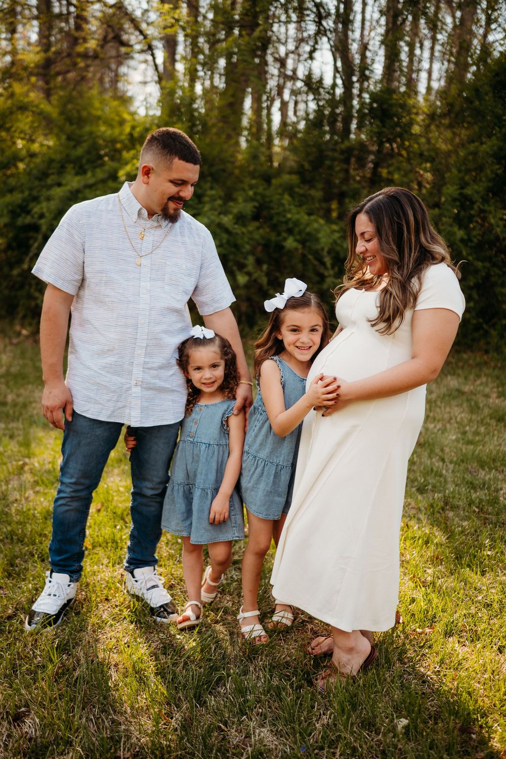 A pregnant woman in white dress poses with her husband and two young daughters in matching blue dresses outdoors.