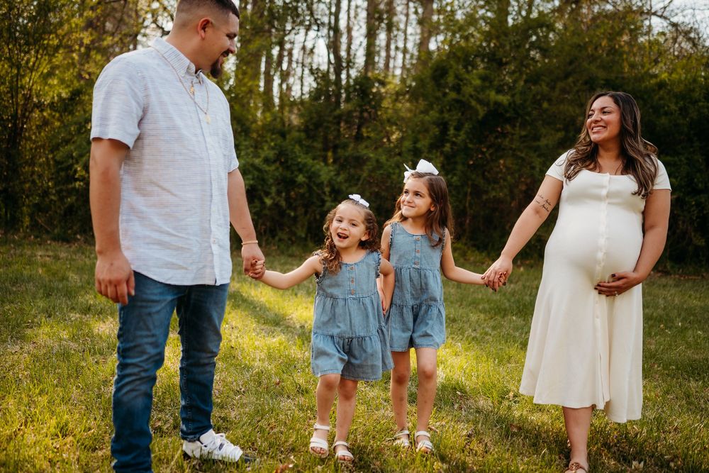 Pregnant woman in white dress holds hands with two girls in denim dresses and husband outdoors.