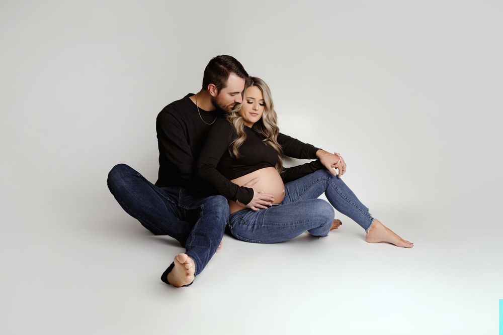 Couple sitting on white studio floor, man in black sweater embracing pregnant woman in jeans, maternity photo session.