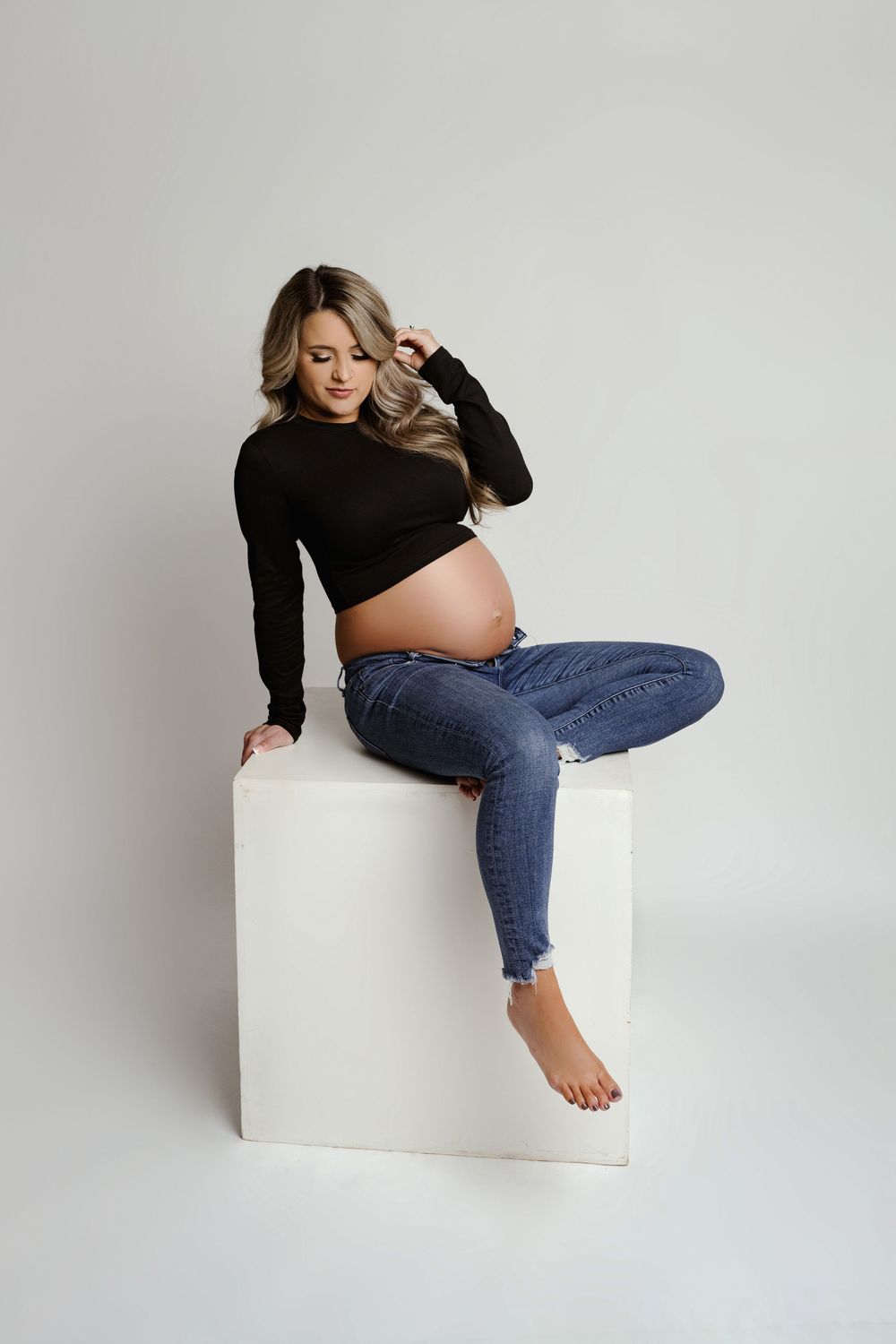 Pregnant woman in black crop top and ripped jeans sitting on white cube, showcasing baby bump in studio maternity photo.