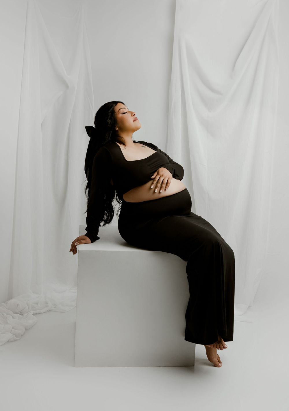 Pregnant woman in black outfit sitting on white cube, posing gracefully against draped white fabric backdrop.
