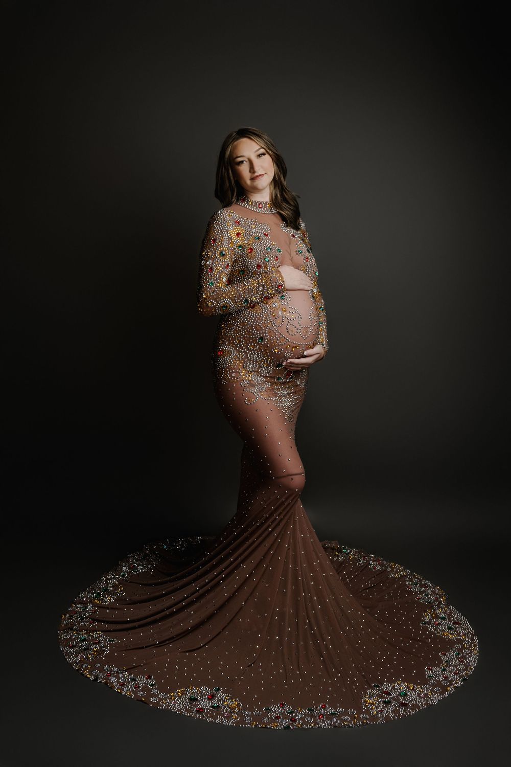 Pregnant woman in elegant floral embellished brown gown with train, cradling baby bump in studio portrait.