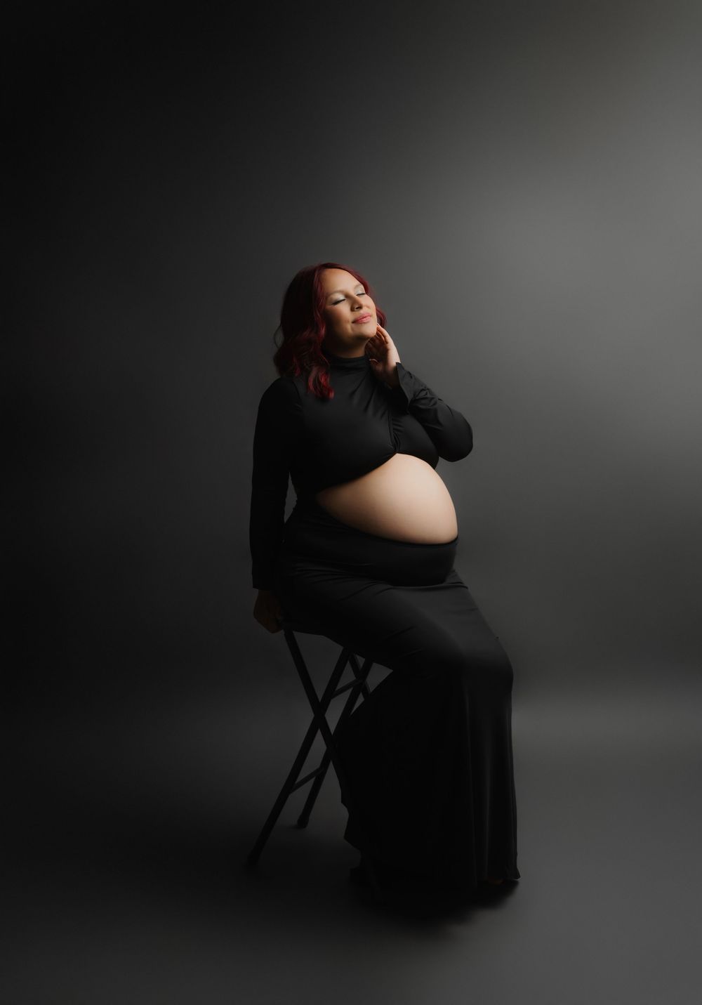 Pregnant woman in black dress sitting on stool, cradling baby bump in studio maternity portrait.