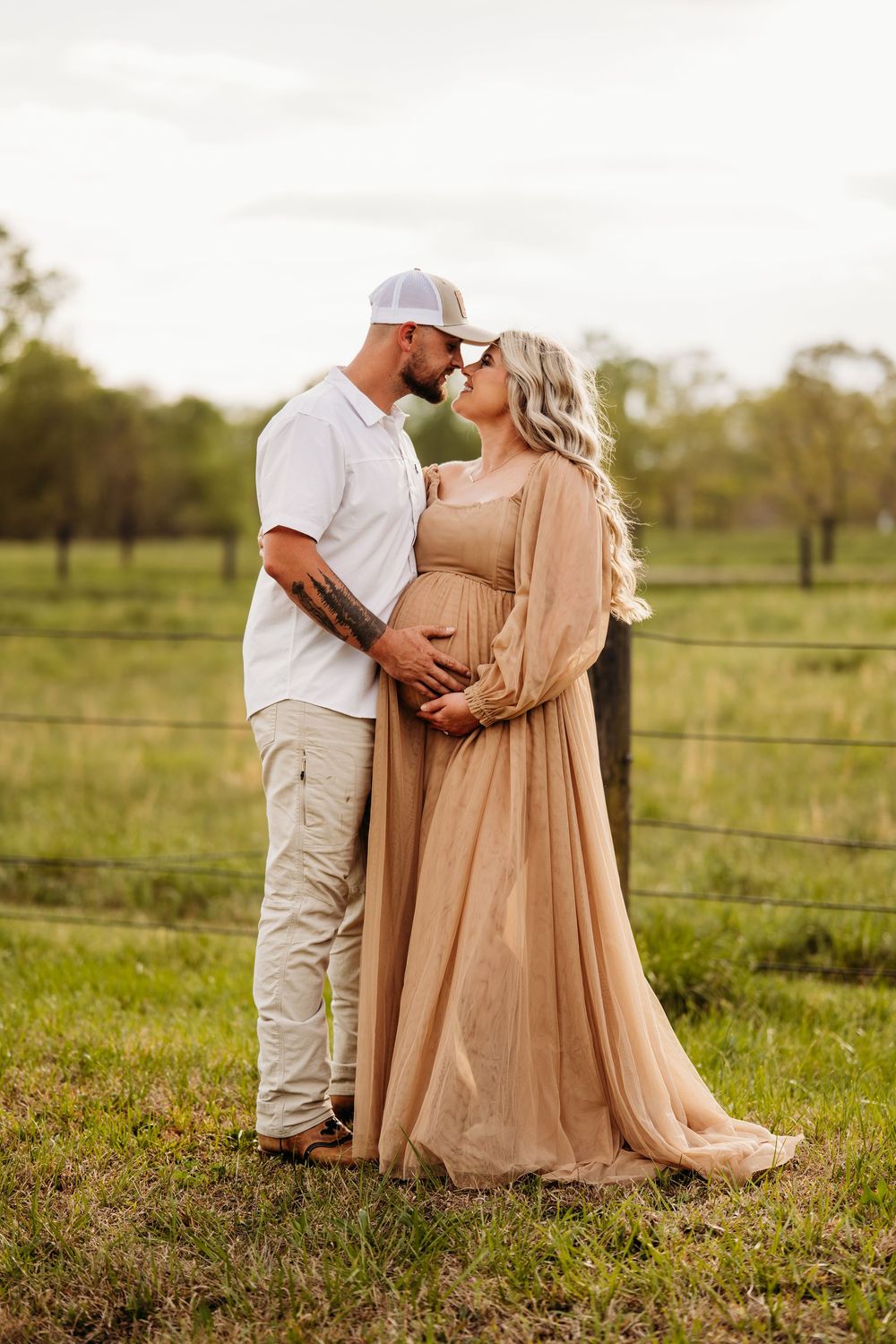 A couple shares a tender moment in a field, the pregnant woman in a flowing champagne gown, her partner in white shirt and cap.