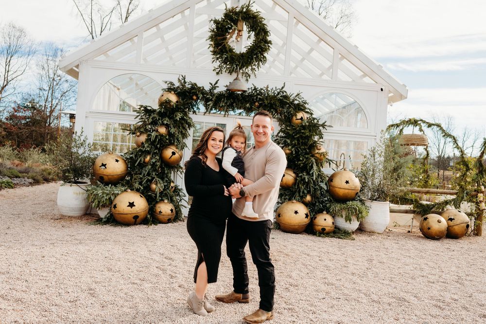 Family of three posing in front of a festive greenhouse decorated with gold ornaments and a holiday wreath.