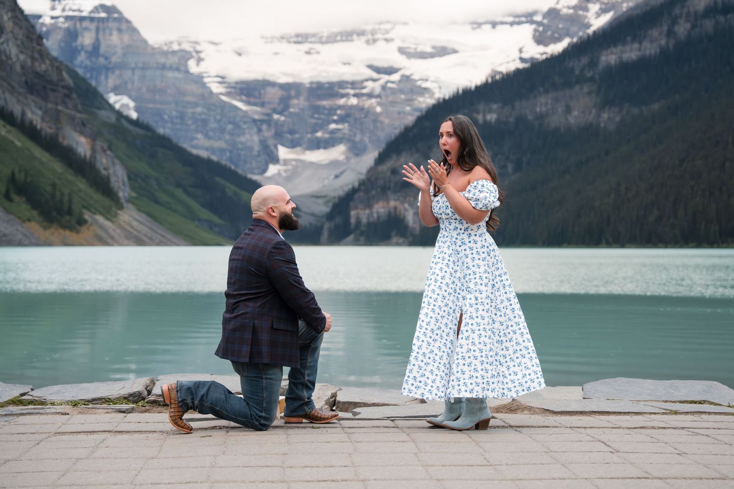 Man proposing on one knee to woman in white dress at Lake Louise, Banff, with turquoise water and mountains behind.