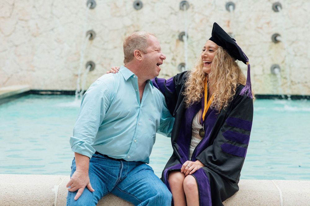 A proud father and his daughter in graduation cap and gown laugh together by a fountain.