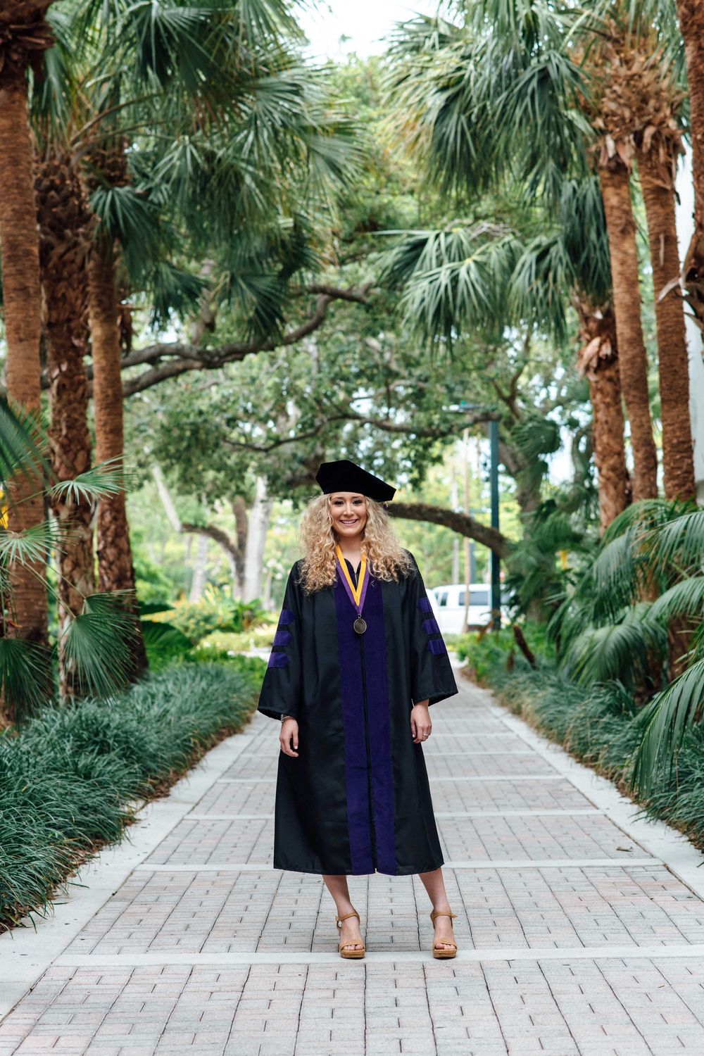 A smiling graduate in cap and gown stands on a palm tree-lined brick pathway.
