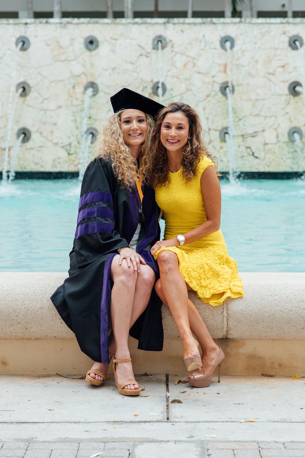 Two women in graduation gown and yellow dress smile together by a fountain.