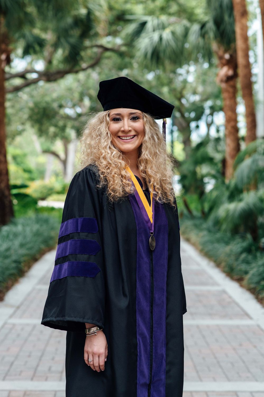Smiling woman in black and purple doctoral graduation cap and gown on campus path.