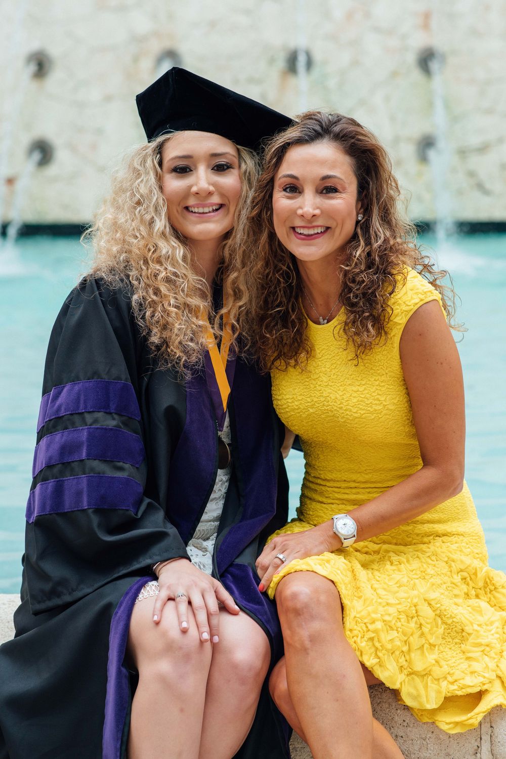 A graduate in cap and gown poses with a woman in a yellow dress at a fountain.