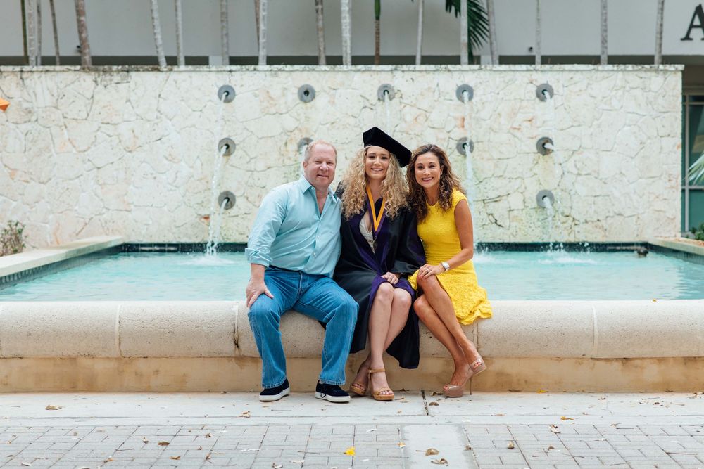 A graduate in cap and gown poses with two proud family members by a fountain outdoors.