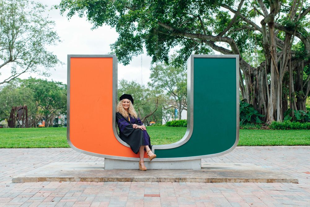 University of Miami graduate in cap and gown sitting on the iconic U sculpture on campus.