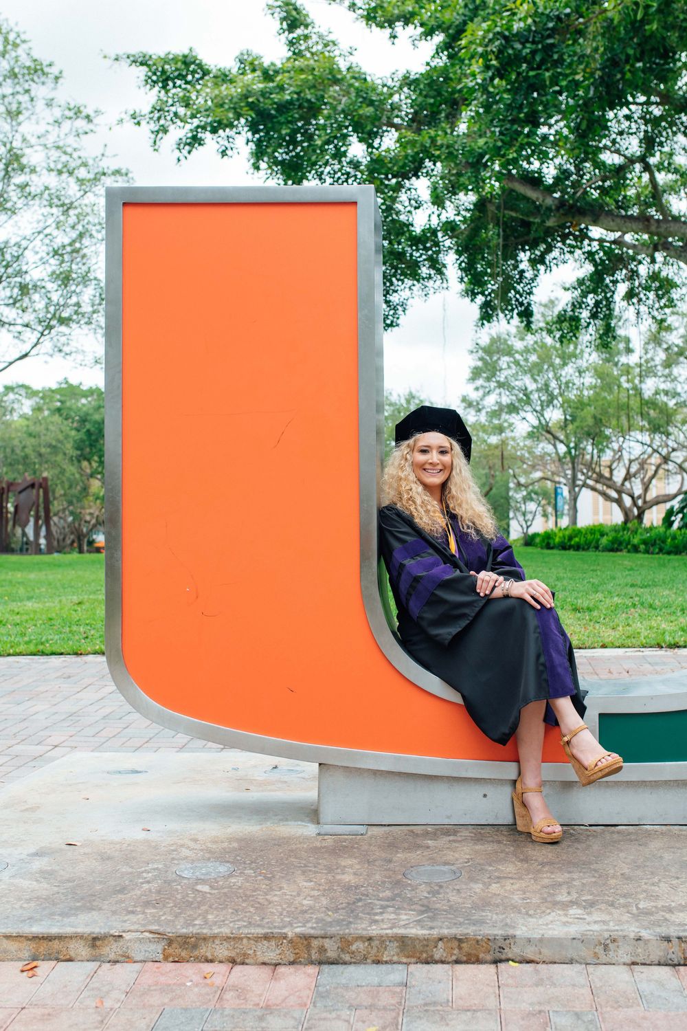 A smiling woman in graduation cap and gown sits beside a large orange University of Miami U sculpture.