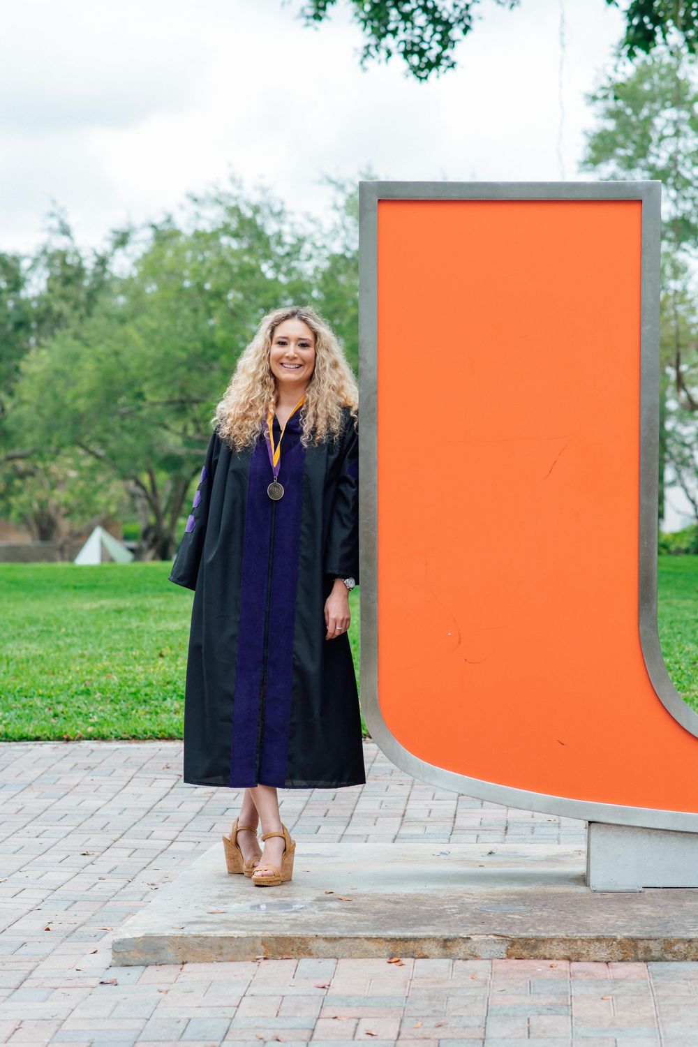 Blonde woman in black graduation gown and medal posing beside an orange university letter monument outdoors.