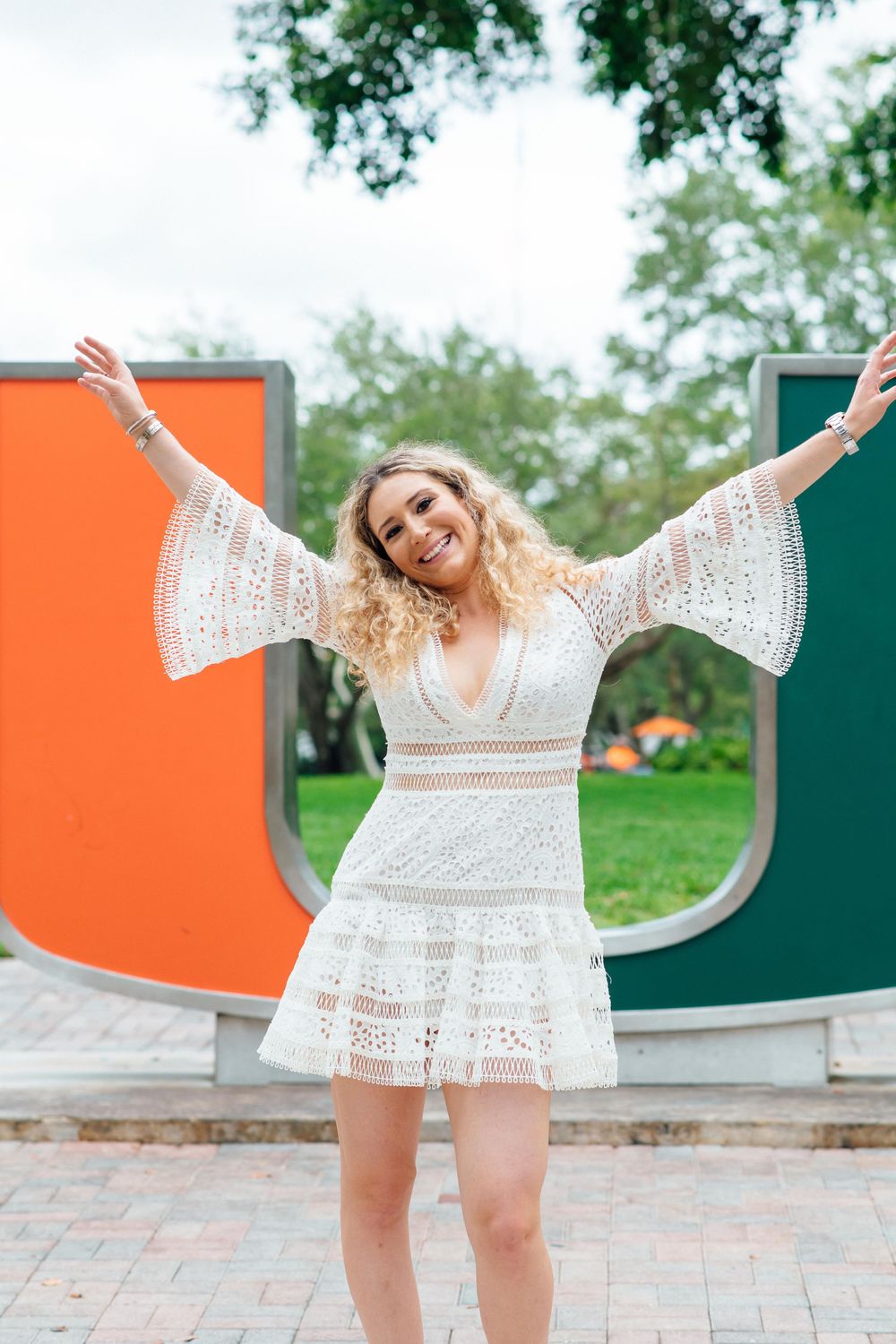 Smiling blonde woman in white lace dress posing with arms wide open in front of University of Miami U sign.