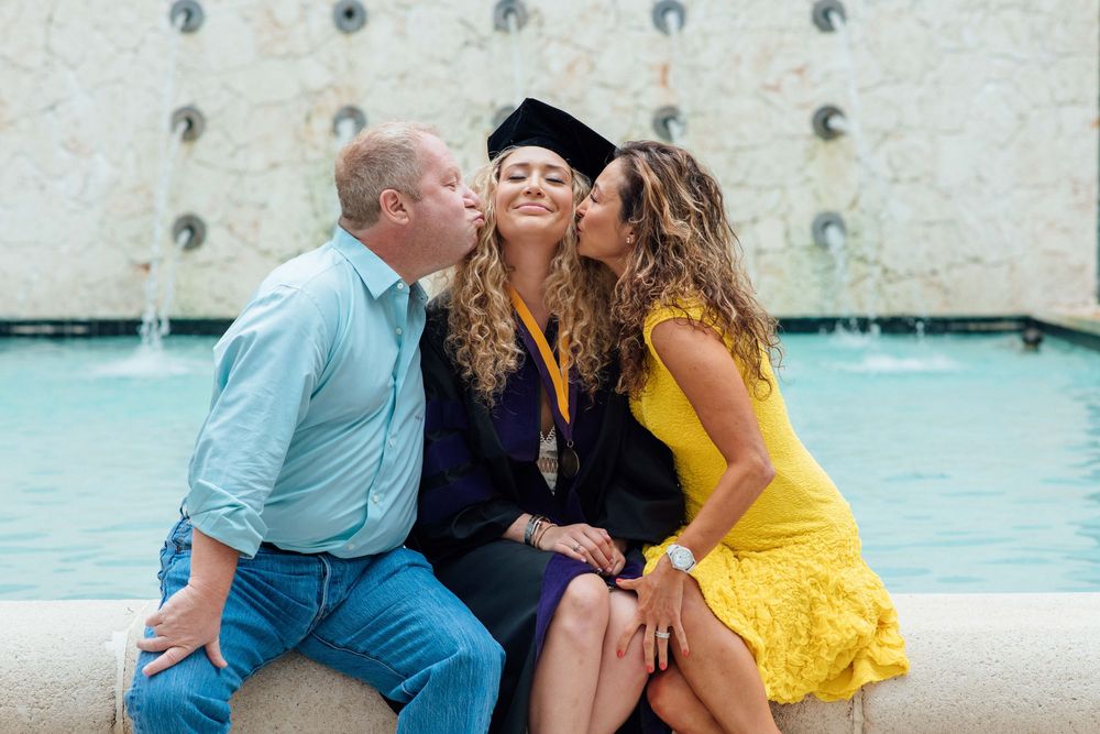 Graduate in cap and gown receives kisses from proud parents seated by a fountain outdoors.