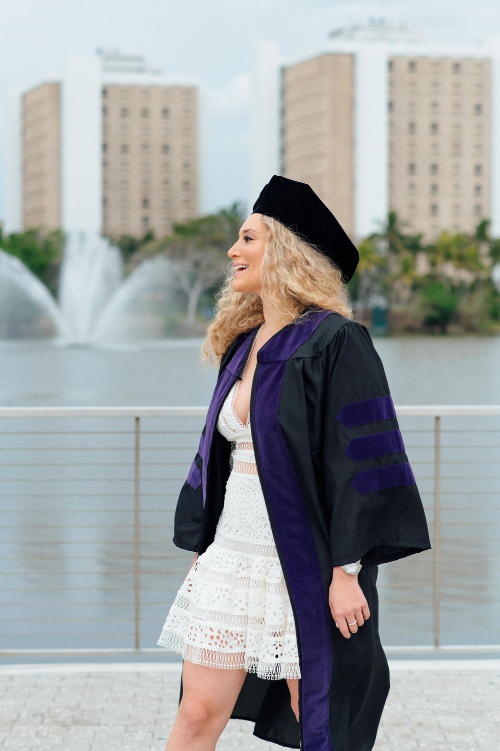 Smiling woman in black graduation gown and cap with purple trim standing near fountain and city buildings.
