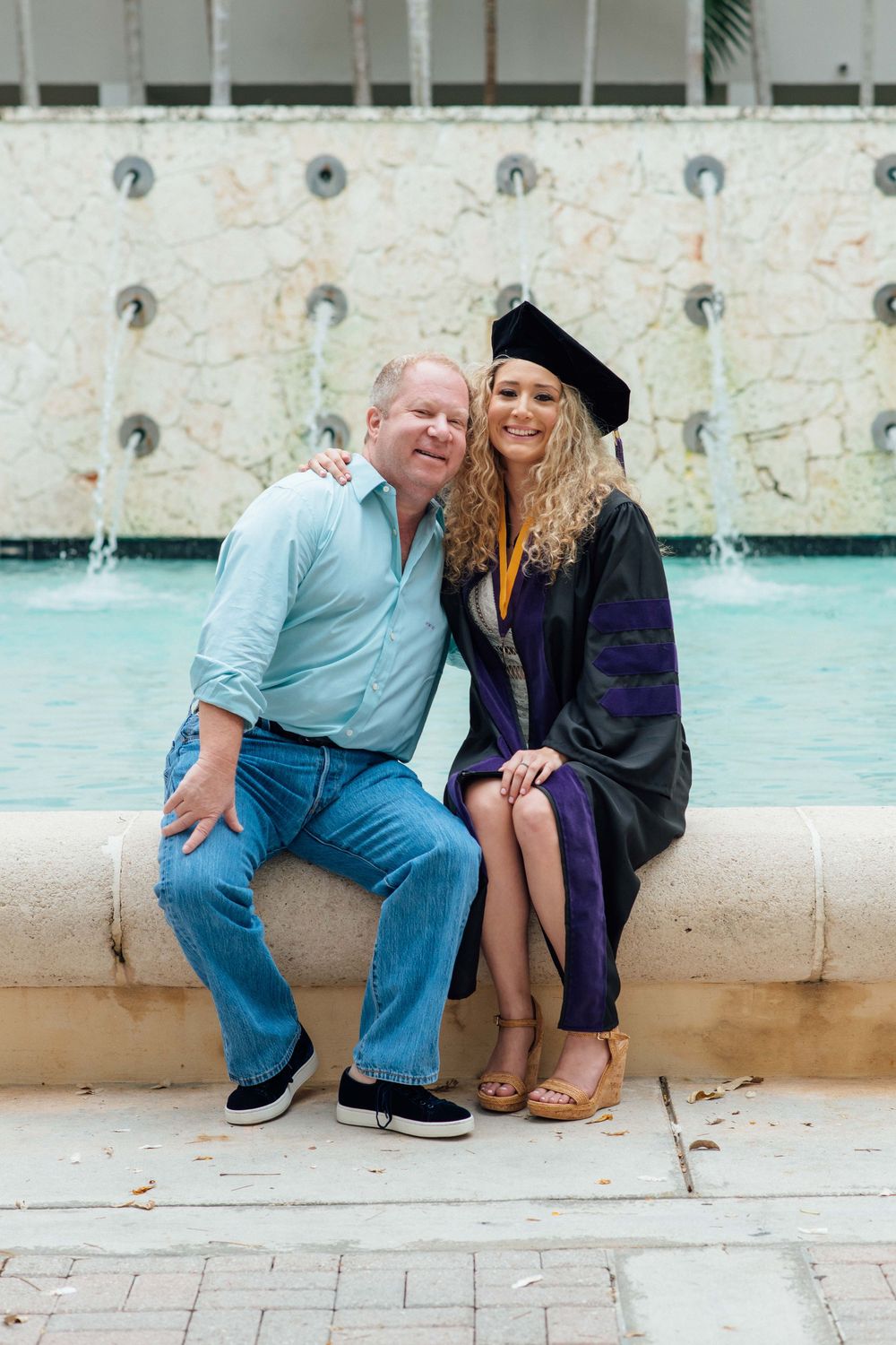 Man and female graduate in cap and gown pose together by a fountain.