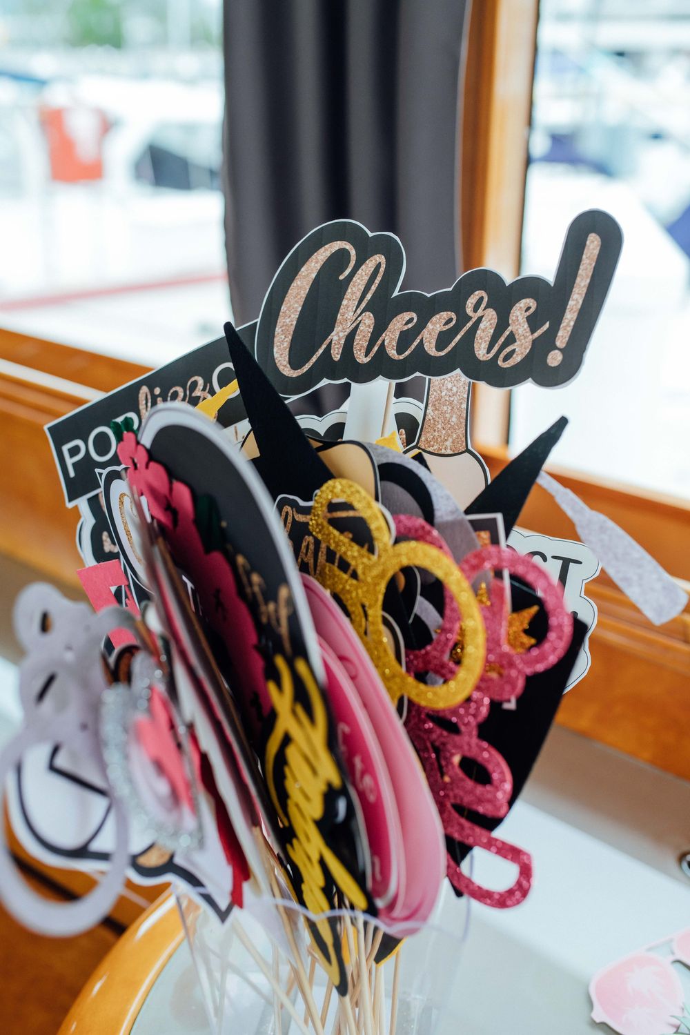 Colorful party photo booth props including Cheers sign, glitter glasses on sticks in a cup.
