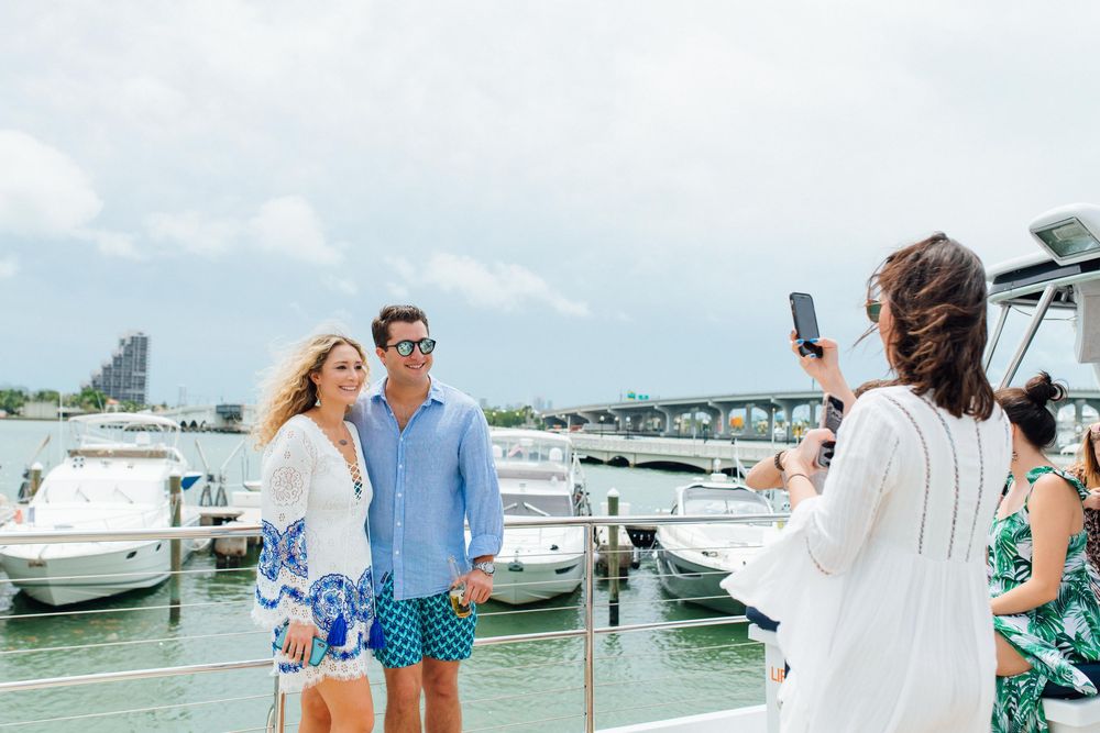 Couple poses for photo on marina dock while friend captures moment on smartphone with boats in background.