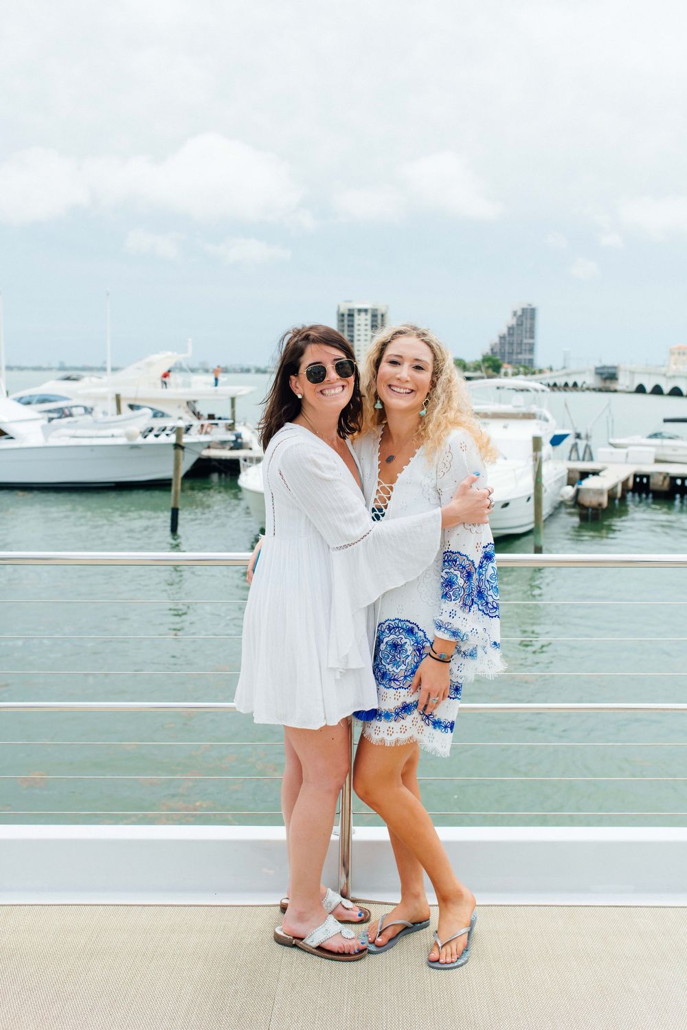 Two smiling women in white and blue outfits posing together on a marina dock with boats in background.
