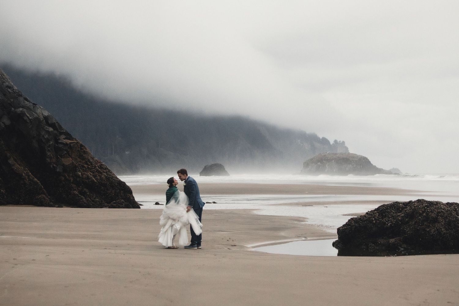 Couple in wedding attire embracing on a misty Oregon coast beach with dramatic sea stacks in the background.