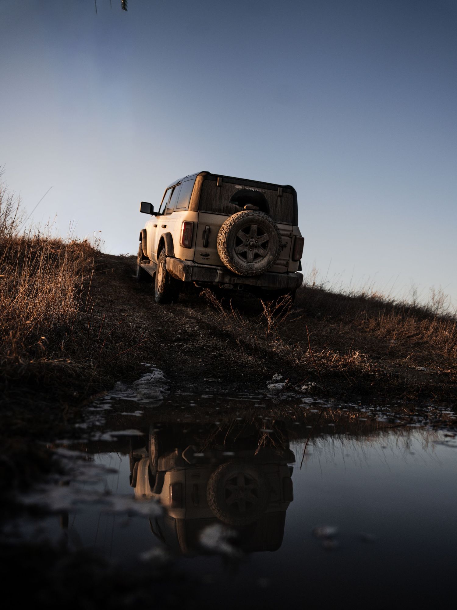 A muddy white Ford Bronco navigates off-road terrain reflected in a puddle at dusk.