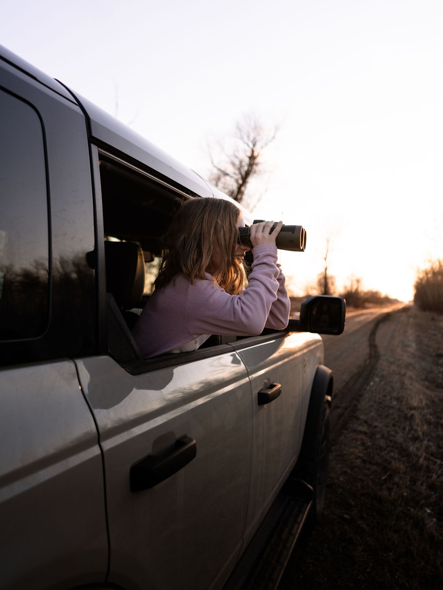 Person leaning out of a white truck window using binoculars on a rural dirt road at sunset.