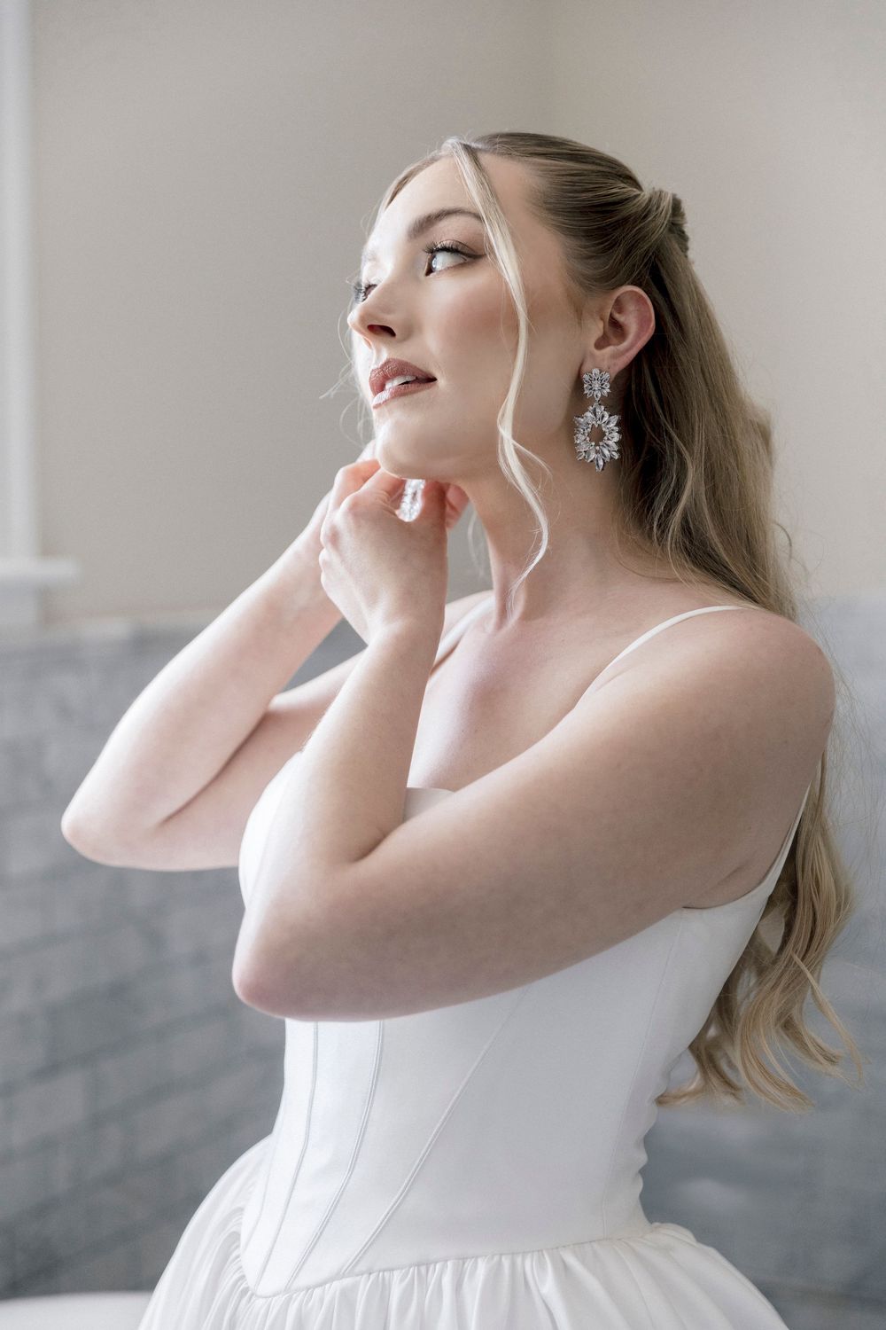 Bride in white corset gown adjusting elegant crystal drop earrings, with soft makeup and blonde hair half-up.