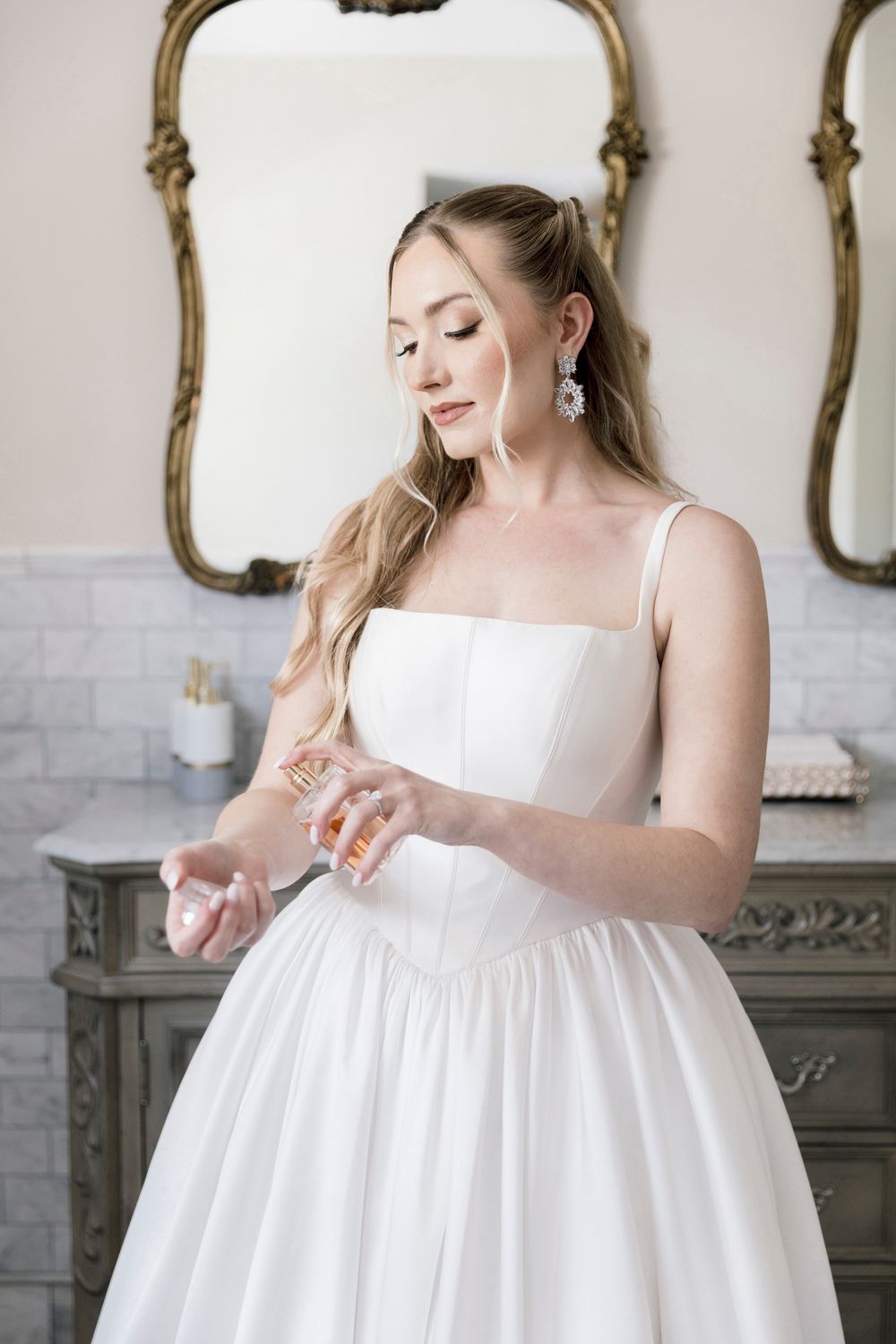 Bride in white ball gown spraying perfume in elegant bathroom with ornate gold mirrors.