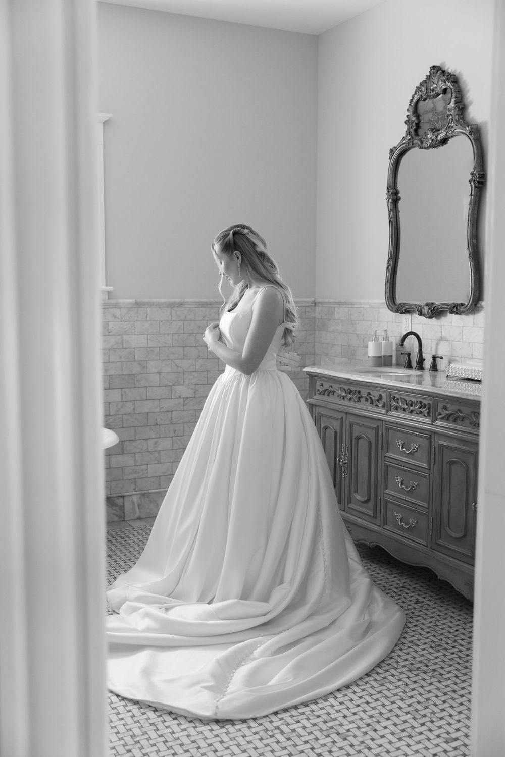 Bride in a flowing white ball gown with train stands in an elegant bathroom with ornate mirror, black and white photo.