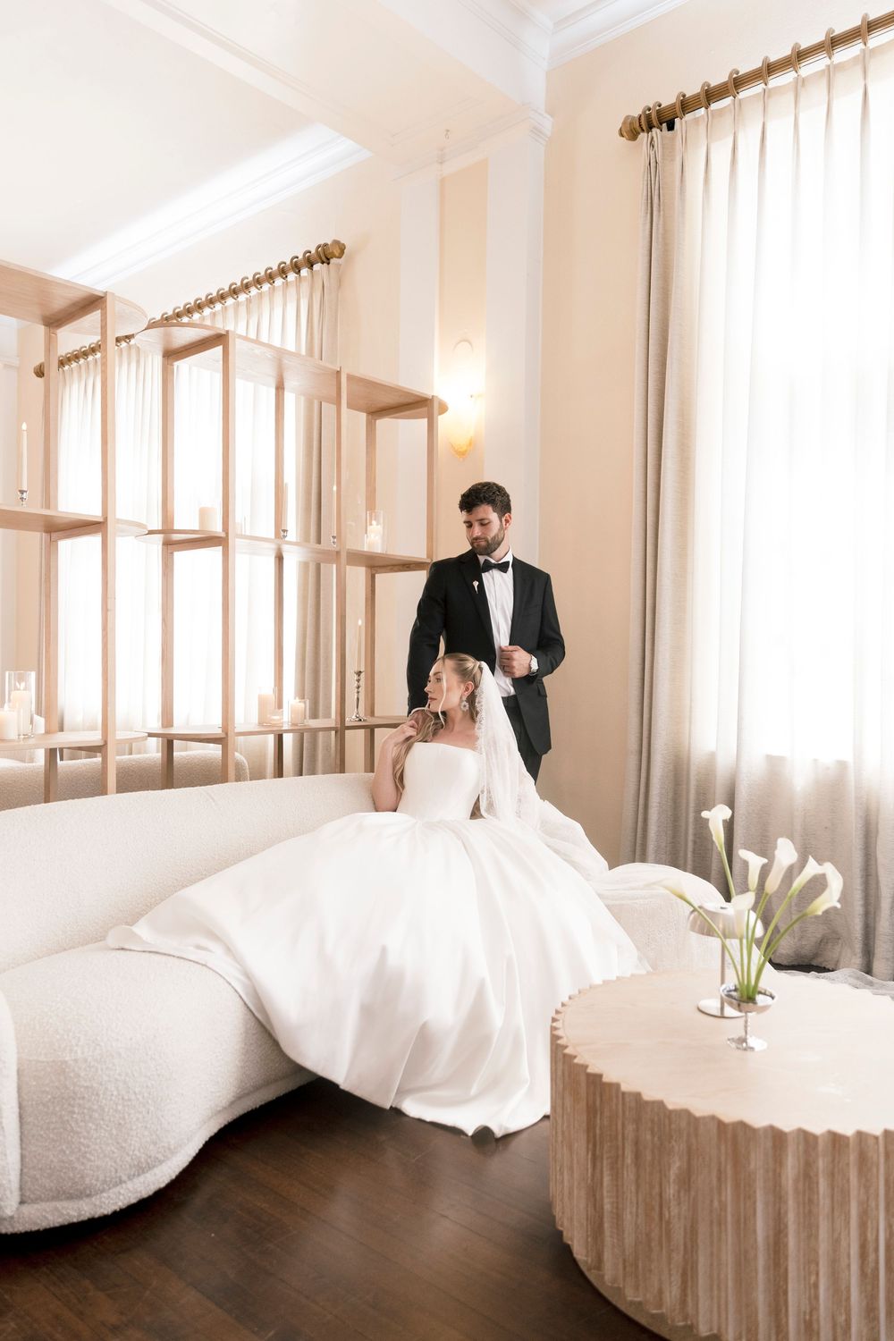 Bride in white gown sits on ivory sofa while groom in black tuxedo stands beside her in elegant room.