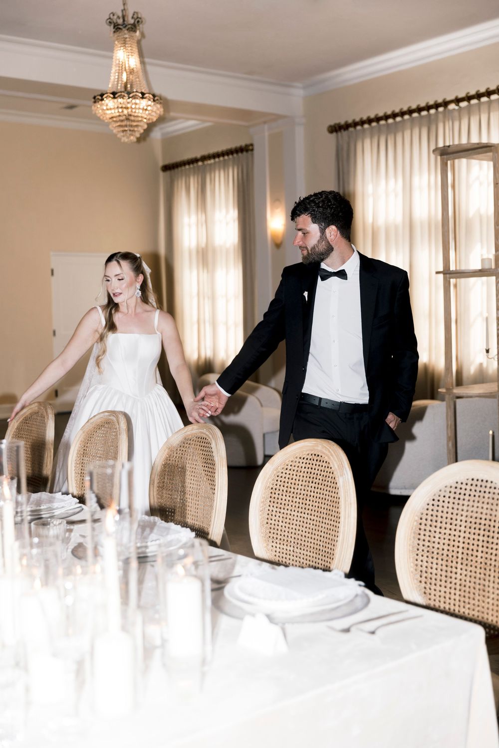 Bride and groom holding hands walking through elegant wedding reception venue with chandelier and rattan chairs.