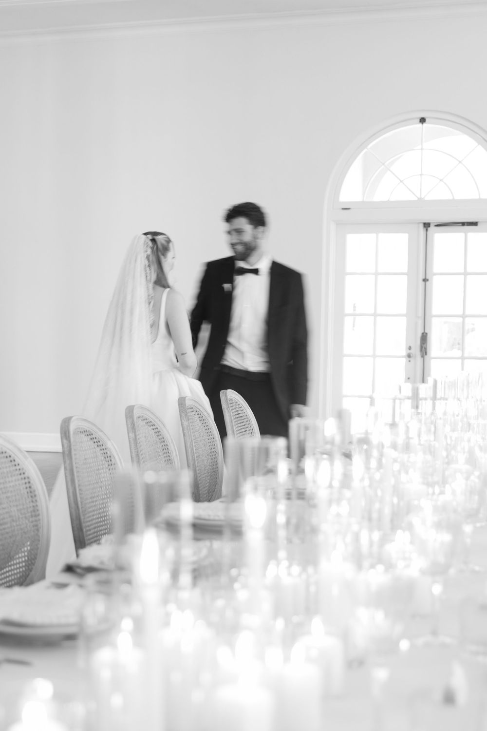 Black and white photo of bride and groom walking through elegant wedding reception hall with candlelit tables.