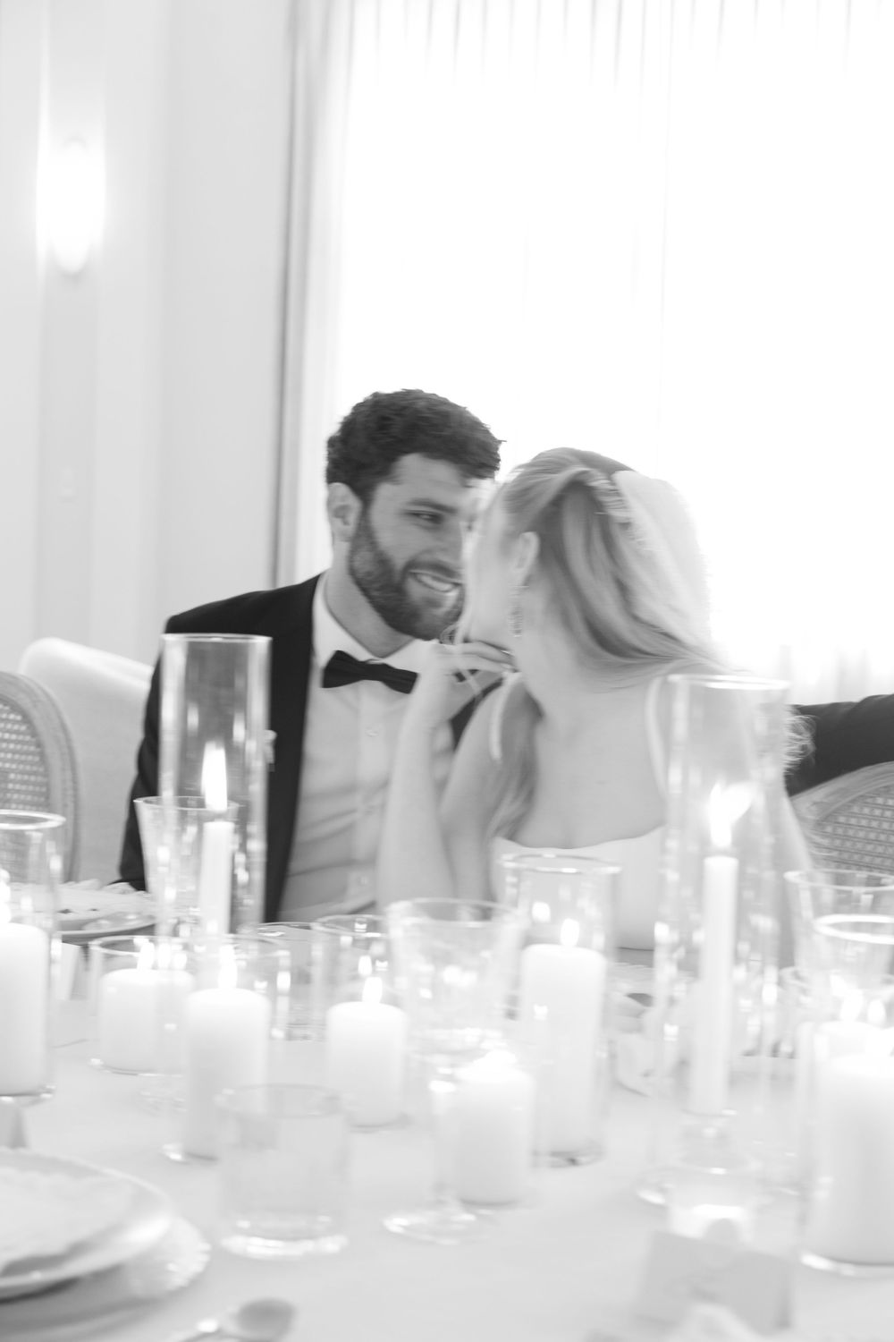 Black and white photo of a smiling groom in a tuxedo sharing an intimate moment with his bride at a candlelit reception table.