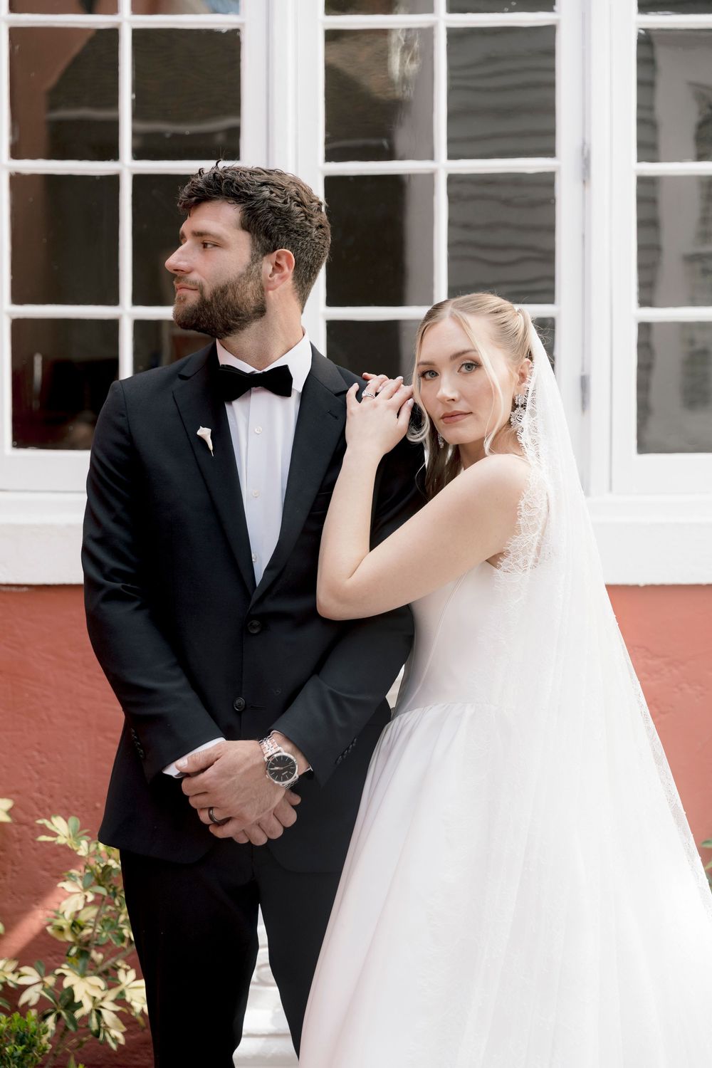 Bride in white tulle gown leans on groom in black tuxedo outdoors near white French windows.