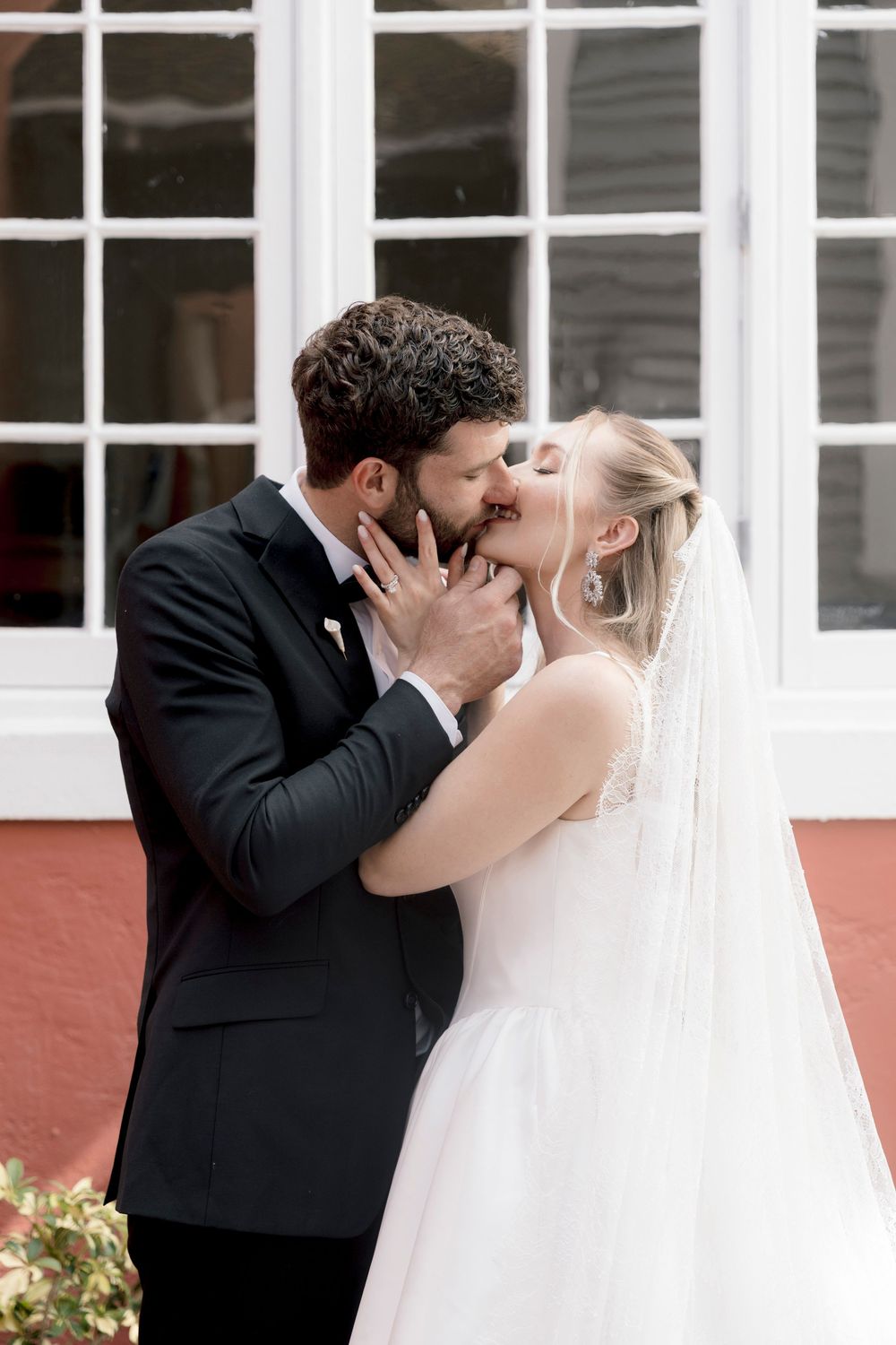 A bride and groom share a romantic kiss outdoors, the bride wearing a white gown and veil, the groom in a black tuxedo.