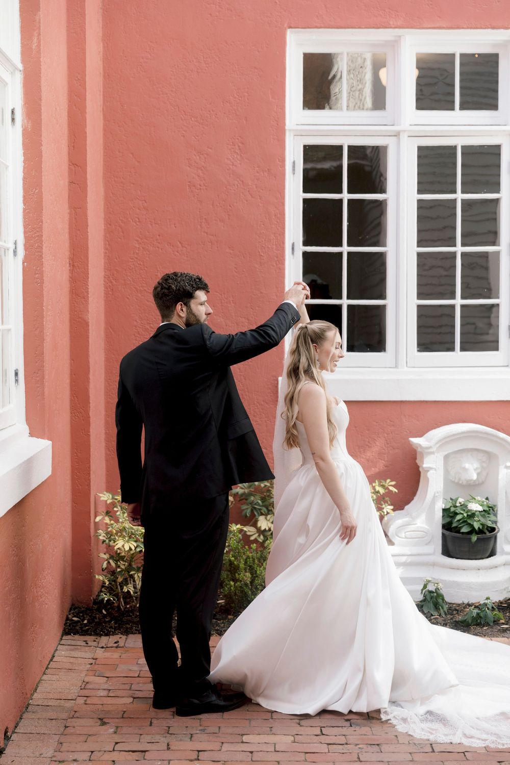 Groom twirling bride in white gown outside pink building with white windows.