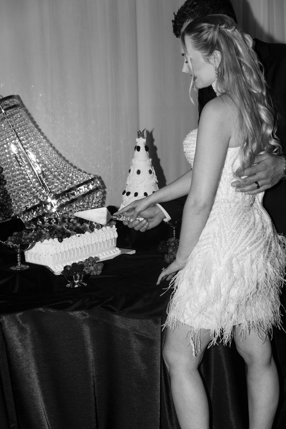 Black and white photo of a bride in a feathered dress cutting a wedding cake with her groom at a reception.