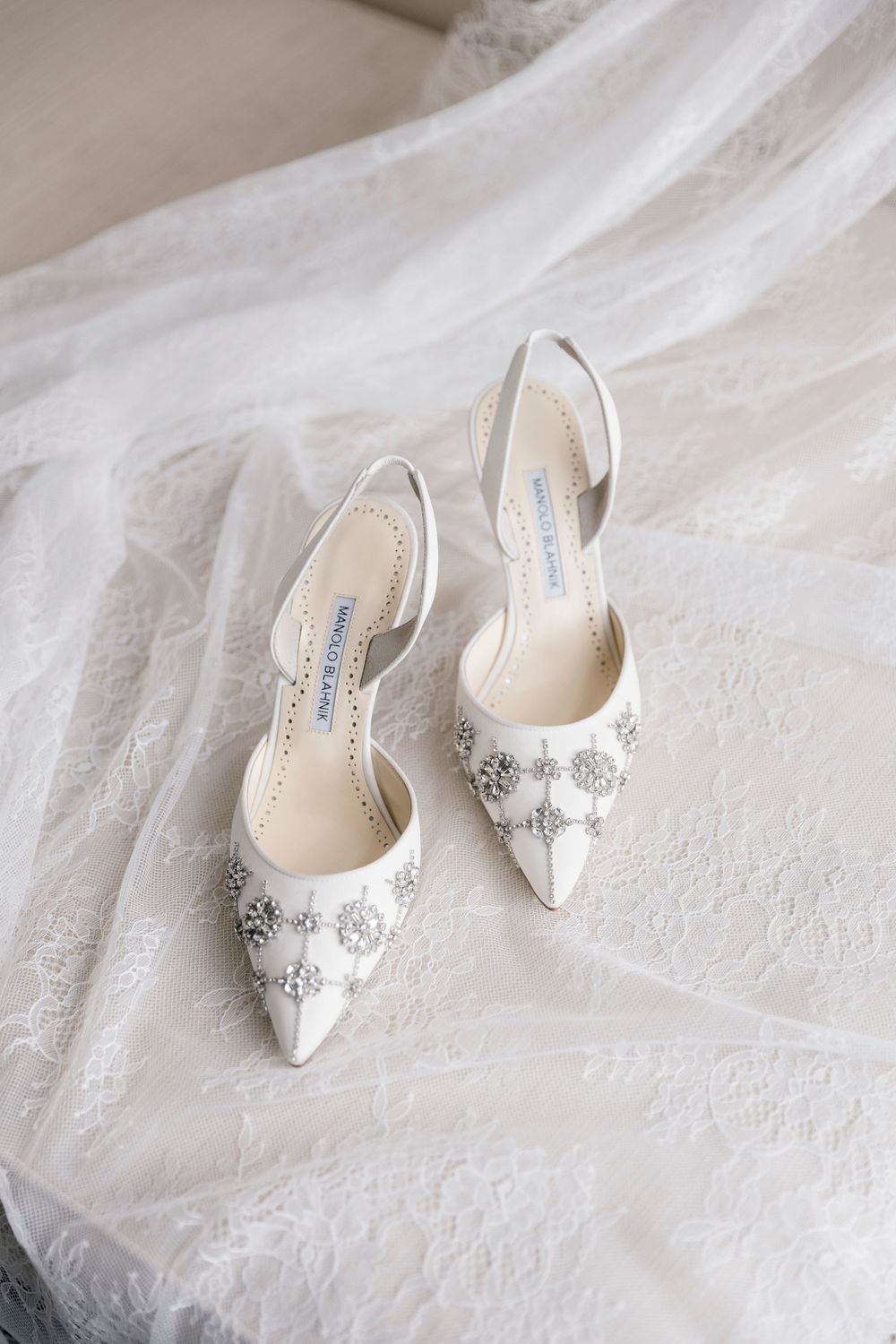 Elegant white Manolo Blahnik bridal heels with crystal embellishments resting on delicate lace wedding veil fabric.