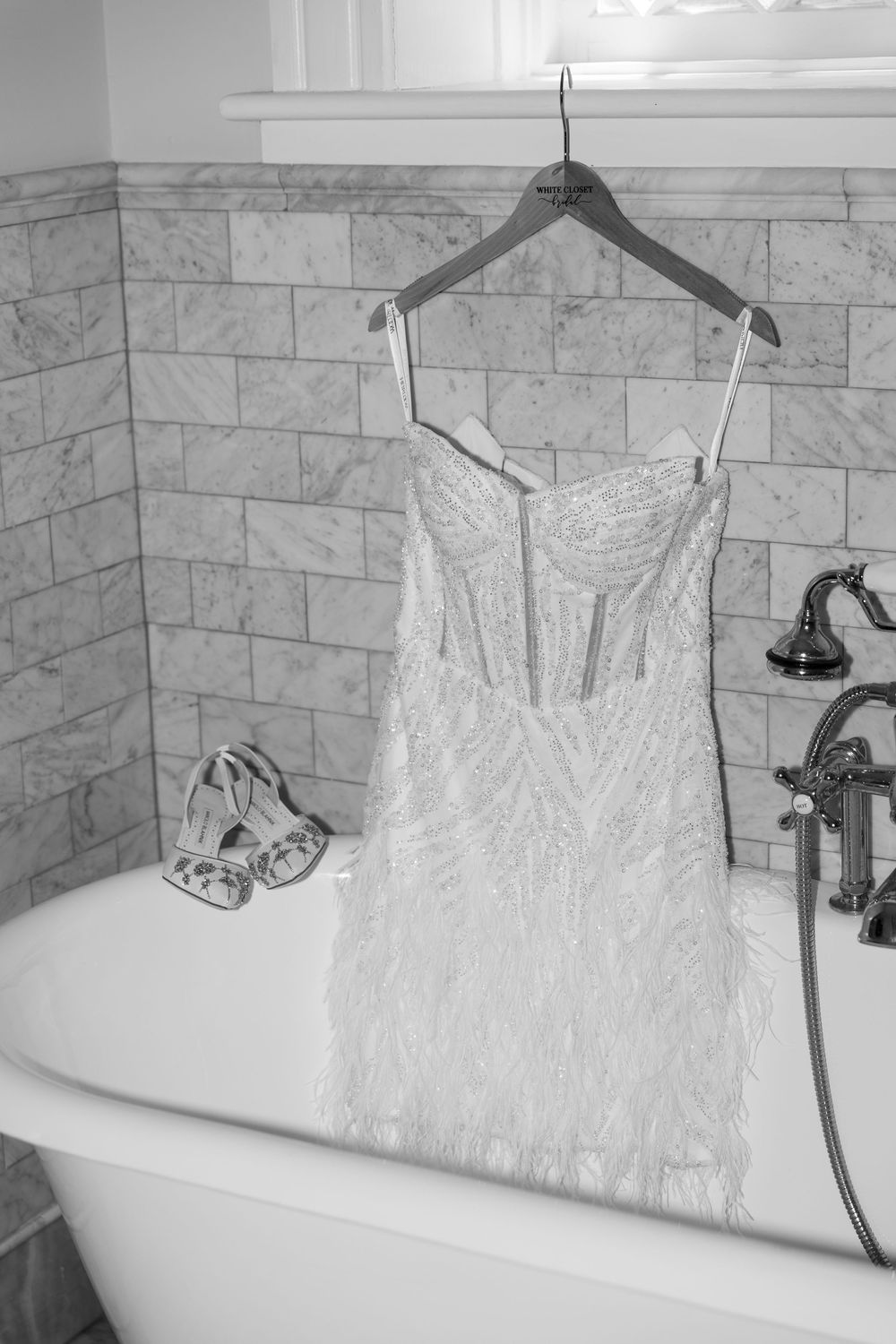 Black and white photo of a beaded wedding dress hanging on a clawfoot bathtub with heels nearby.