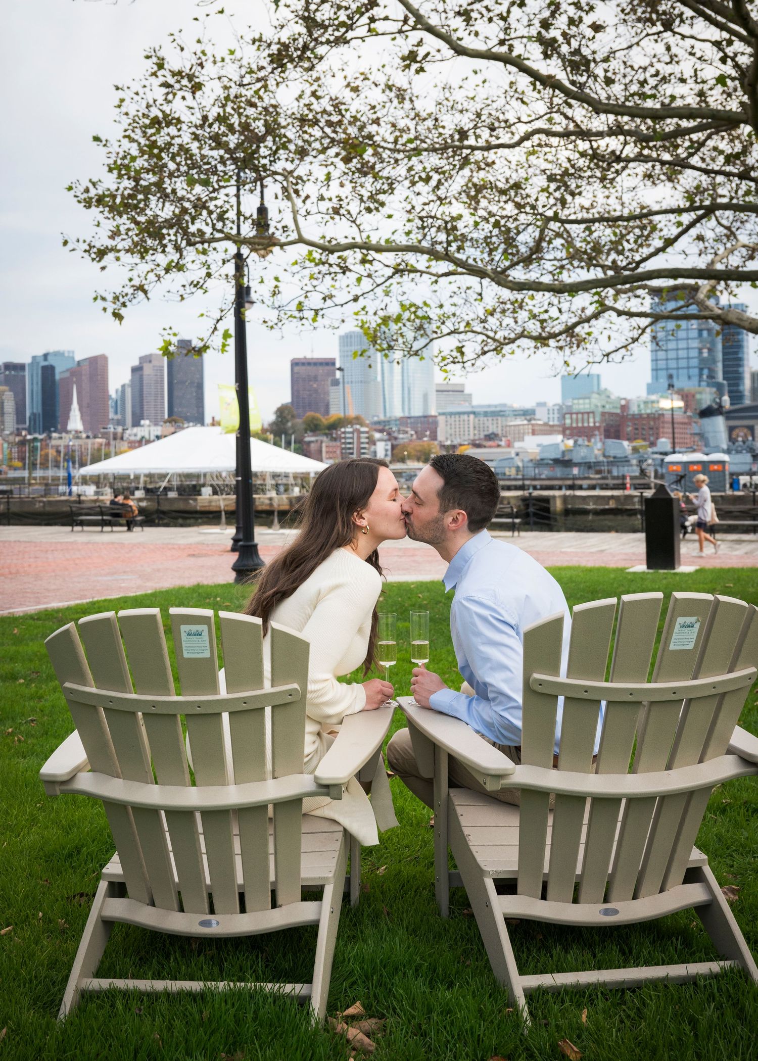 Couple kissing in Adirondack chairs on waterfront lawn with Boston city skyline in background.