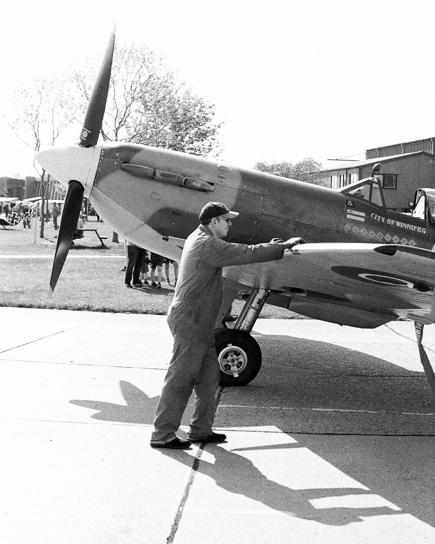 A mechanic in coveralls inspects a vintage WWII Spitfire fighter aircraft on a tarmac in a black and white photo.