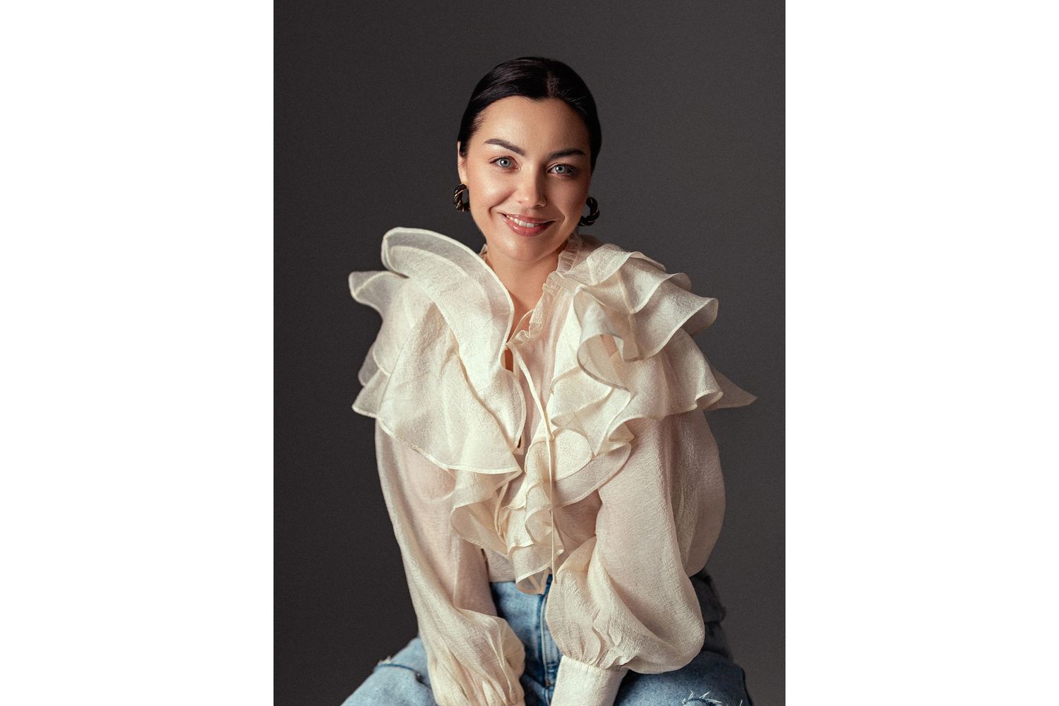 Woman in cream ruffled blouse sits smiling confidently in stylish portrait against dark gray background.