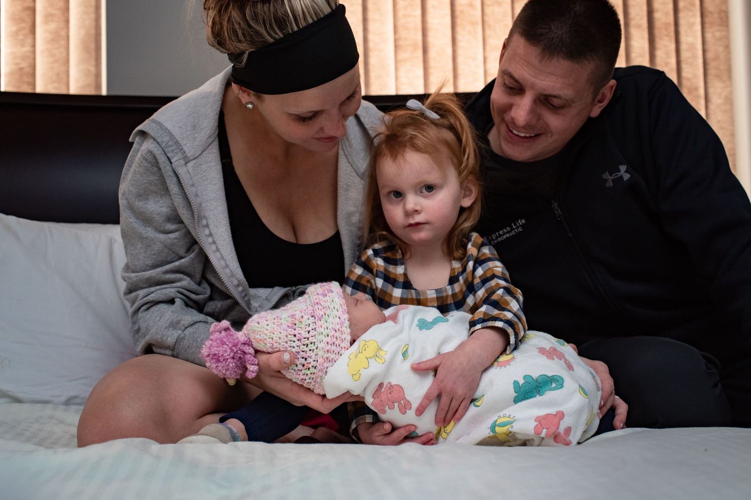 A family sits together on a bed bonding with their newborn wrapped in patterned blankets.