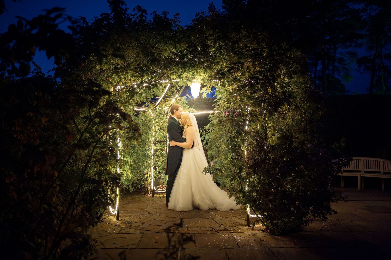 Bride and groom kissing under a lit garden arch at night, surrounded by lush greenery in a romantic outdoor setting.