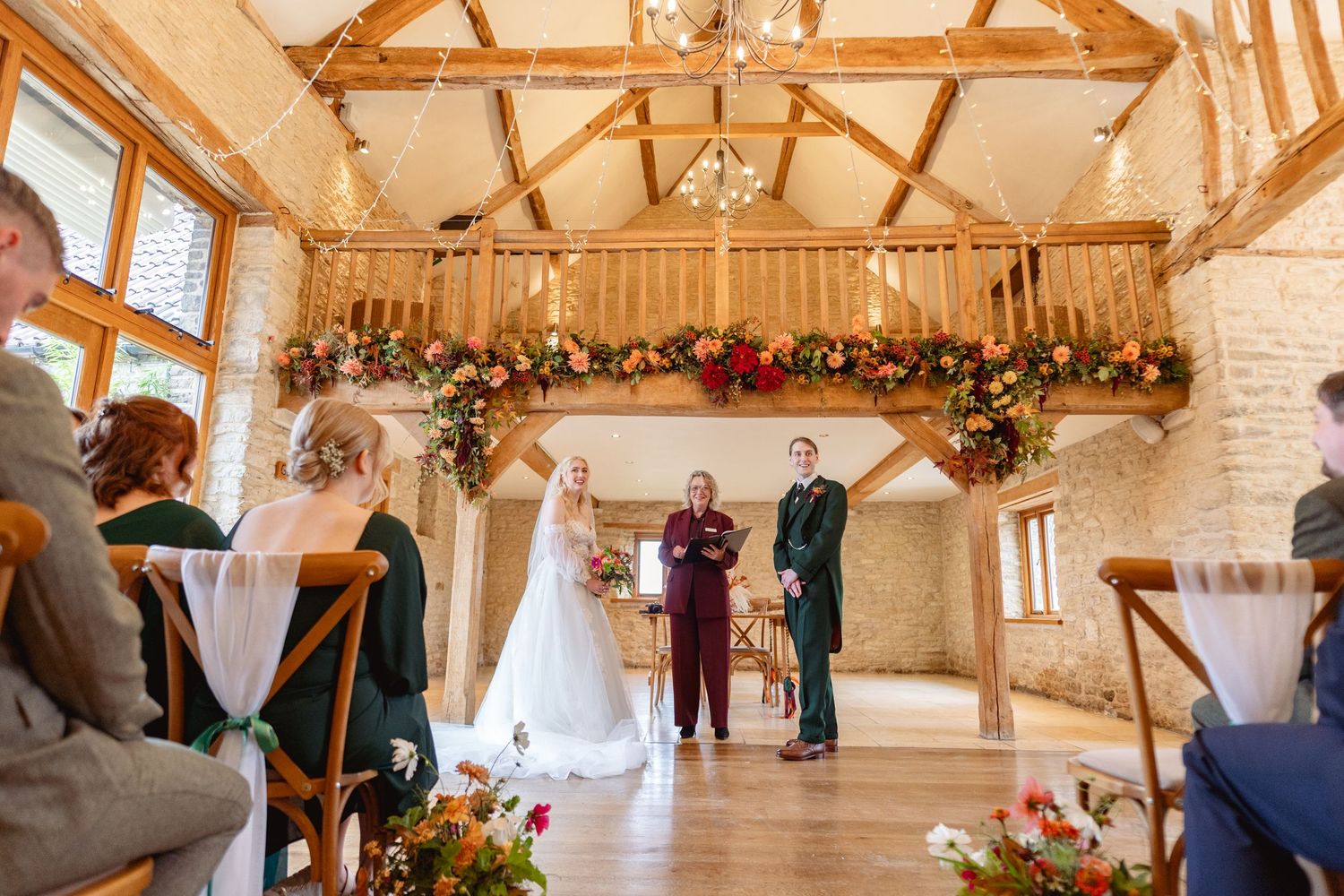 Rustic barn wedding ceremony with floral balcony, bride and groom at altar, exposed wooden beams, and seated guests.