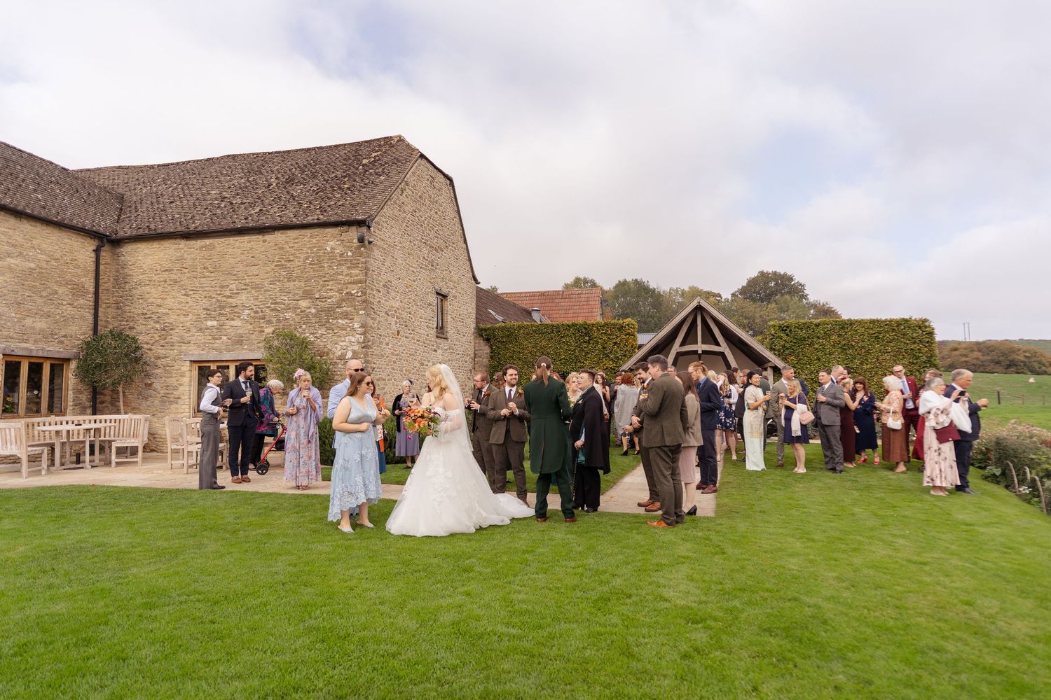 Outdoor wedding ceremony on lush green lawn beside rustic stone barn venue with guests gathered around the couple.